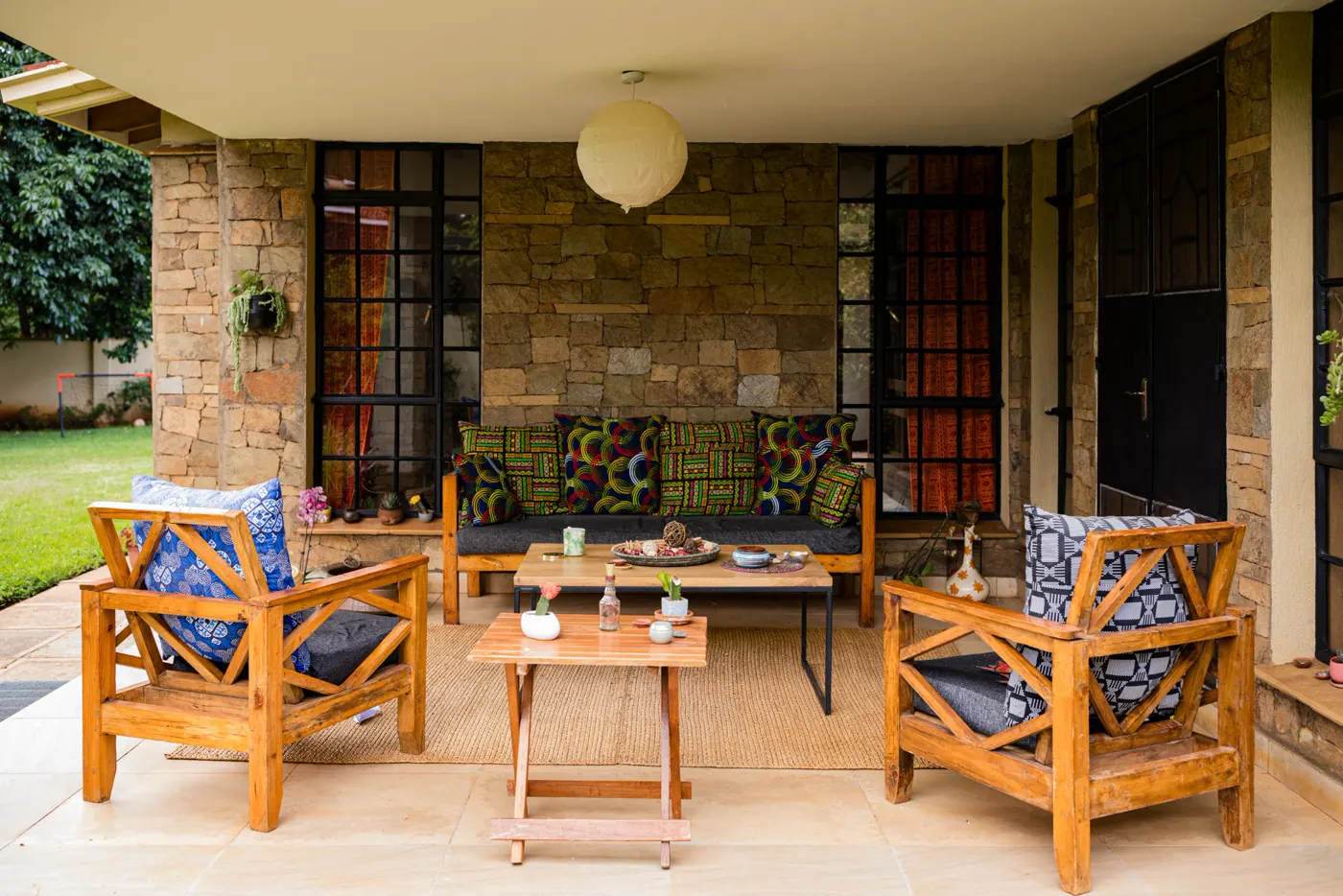 Terrasse couverte aux murs en pierre, mobilier en bois et coussins en tissu wax